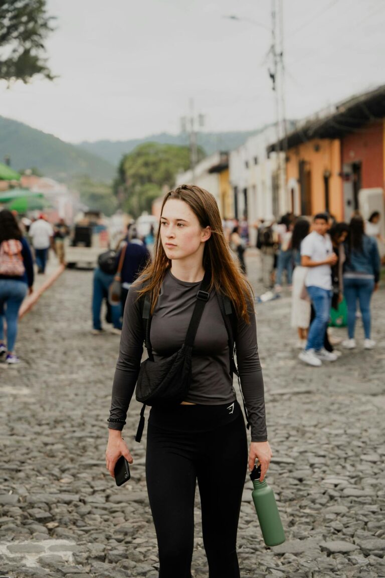 A young woman walks through a lively cobblestone street market, surrounded by people and colorful buildings.